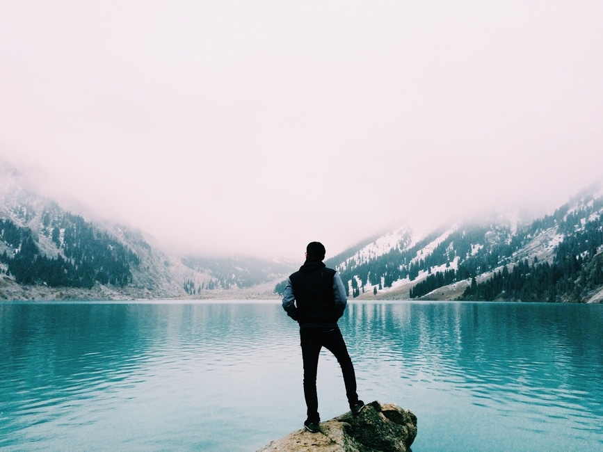 A person standing at the edge of a turquoise mountain lake, looking towards misty peaks